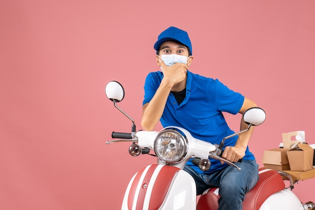 Vista frontal del mensajero con máscara médica con sombrero sentado en scooter y mirando algo con expresión facial sorprendida sobre fondo melocotón pastel