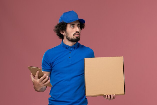 Vista frontal del mensajero masculino con gorra uniforme azul con bloc de notas y caja de comida en sus manos en la pared rosa