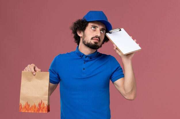 Vista frontal del mensajero masculino en gorra uniforme azul con bloc de notas y paquete de comida de entrega en sus manos pensando en la pared rosa