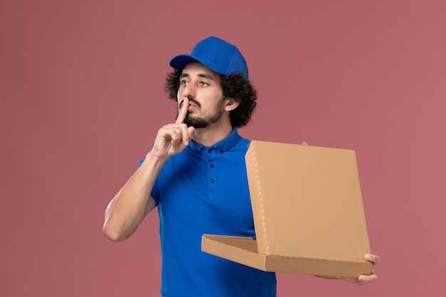 Vista frontal del mensajero masculino con gorra de uniforme azul con caja de comida abierta en sus manos en la pared rosa claro