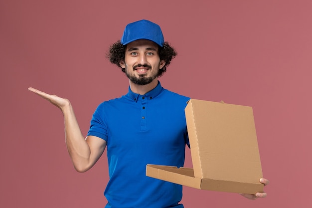 Vista frontal del mensajero masculino con gorra de uniforme azul con caja de comida abierta en sus manos en la pared rosa claro