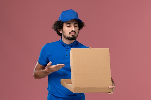 Vista frontal del mensajero masculino con gorra de uniforme azul con caja de comida abierta en sus manos que huele en la pared rosa claro