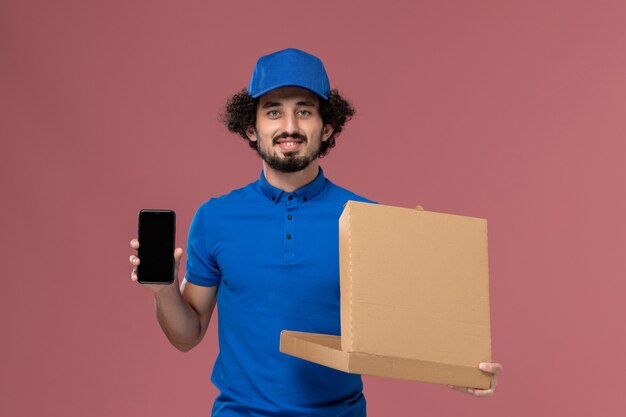 Vista frontal del mensajero masculino con gorra de uniforme azul con caja de comida abierta y teléfono en sus manos en la pared rosa claro