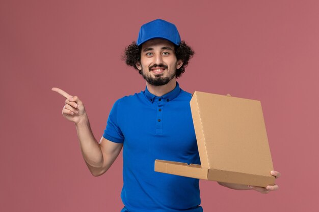 Vista frontal del mensajero masculino con gorra de uniforme azul con caja de comida en sus manos abriéndola en la pared rosa claro