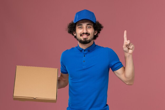 Vista frontal del mensajero masculino con gorra de uniforme azul con caja de comida en sus manos en la pared rosa claro