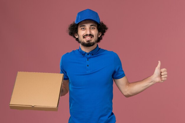 Vista frontal del mensajero masculino con gorra de uniforme azul con caja de comida en sus manos en la pared rosa claro