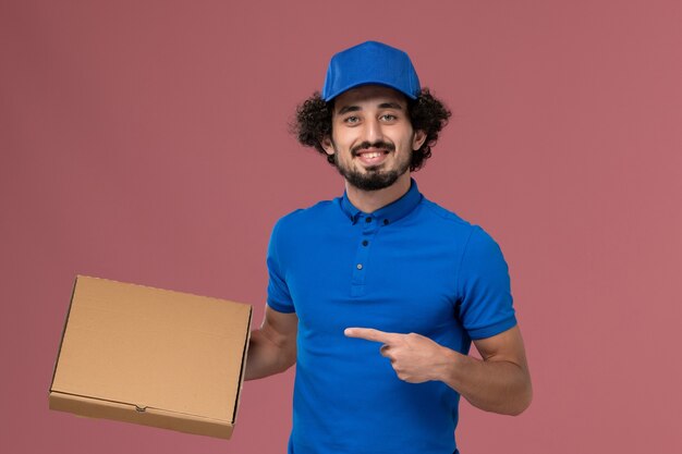 Vista frontal del mensajero masculino con gorra de uniforme azul con caja de comida en sus manos en la pared rosa claro
