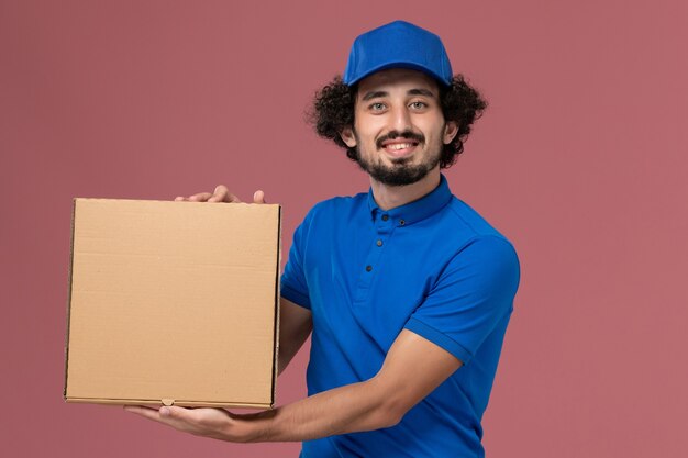 Vista frontal del mensajero masculino con gorra de uniforme azul con caja de comida en sus manos sonriendo en la pared de color rosa claro