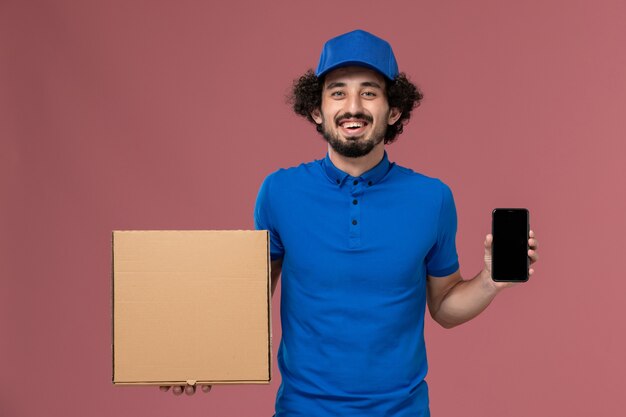 Vista frontal del mensajero masculino con gorra de uniforme azul con caja de comida y teléfono en sus manos en la pared rosa claro