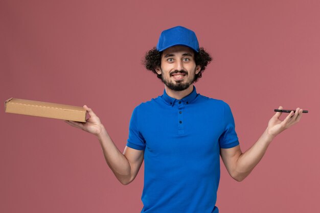 Vista frontal del mensajero masculino con gorra de uniforme azul con caja de comida y teléfono en sus manos en la pared rosa