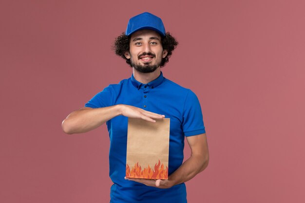Vista frontal del mensajero masculino con gorra uniforme azul con paquete de comida de papel de entrega en sus manos en la pared rosa claro