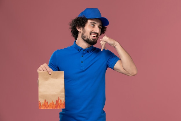 Vista frontal del mensajero masculino con gorra uniforme azul con paquete de comida de papel de entrega en sus manos en la pared rosa claro