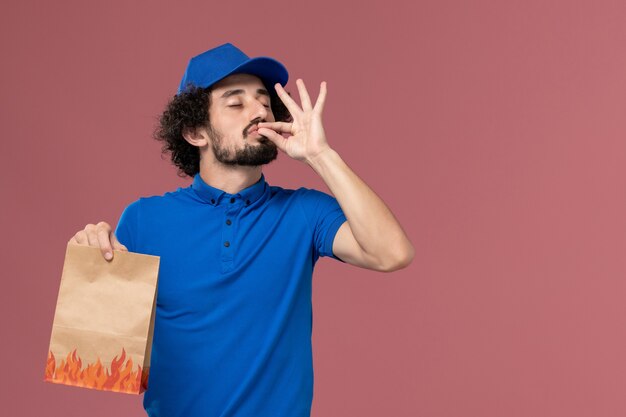 Vista frontal del mensajero masculino con gorra uniforme azul con paquete de comida de papel de entrega en sus manos en la pared rosa claro