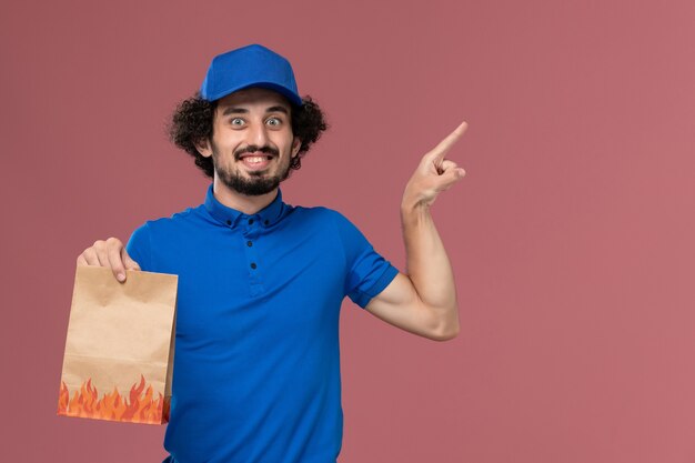 Vista frontal del mensajero masculino con gorra uniforme azul con paquete de comida de papel de entrega en sus manos en la pared rosa claro