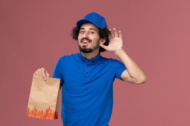 Vista frontal del mensajero masculino con gorra de uniforme azul con paquete de comida de papel de entrega en sus manos tratando de escuchar en la pared rosa claro