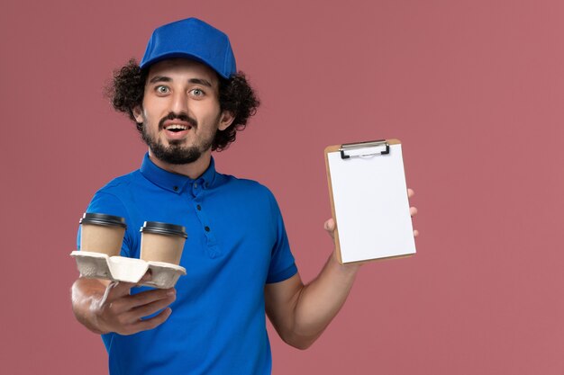 Vista frontal del mensajero masculino en gorra uniforme azul con tazas de café de entrega y bloc de notas en sus manos en la pared rosa