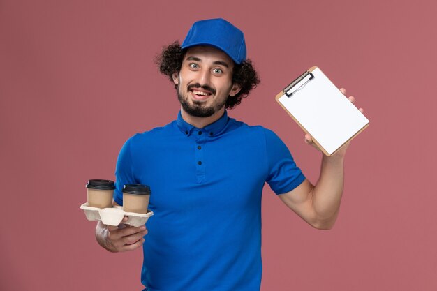 Vista frontal del mensajero masculino en gorra uniforme azul con tazas de café de entrega y bloc de notas en sus manos en la pared rosa