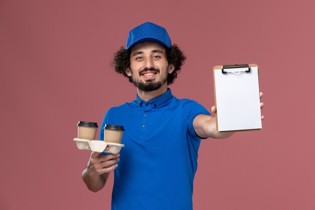 Vista frontal del mensajero masculino en gorra uniforme azul con tazas de café de entrega y bloc de notas en sus manos en la pared rosa