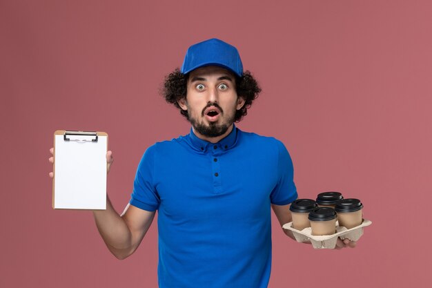 Vista frontal del mensajero masculino en gorra uniforme azul con tazas de café de entrega y bloc de notas en sus manos en la pared rosa