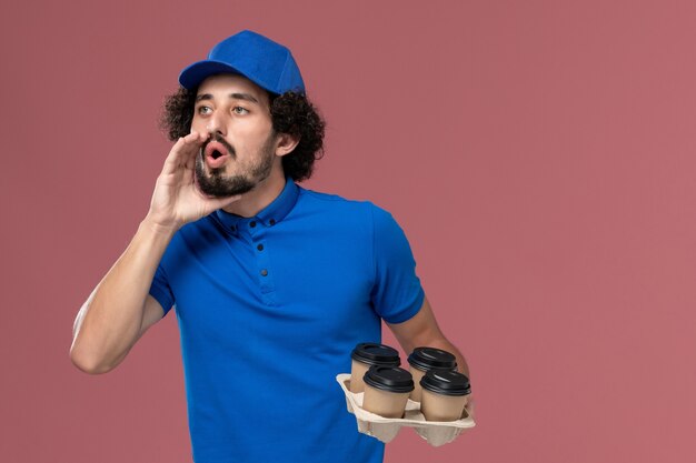 Vista frontal del mensajero masculino con gorra de uniforme azul con tazas de café de entrega en sus manos llamando a la pared rosa claro