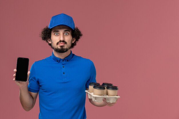 Vista frontal del mensajero masculino con gorra de uniforme azul con tazas de café de entrega en sus manos y su teléfono de trabajo en la pared rosa