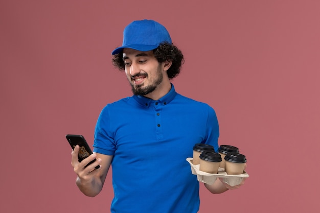 Foto gratuita vista frontal del mensajero masculino con gorra de uniforme azul con tazas de café de entrega en sus manos y su teléfono de trabajo en la pared rosa