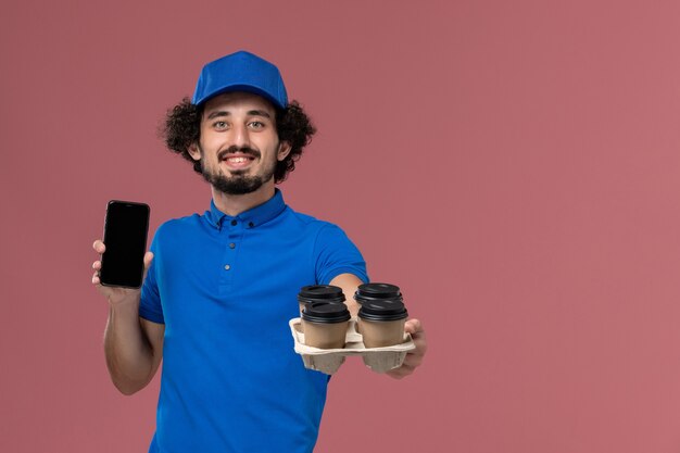 Vista frontal del mensajero masculino con gorra de uniforme azul con tazas de café de entrega en sus manos y su teléfono de trabajo en la pared rosa