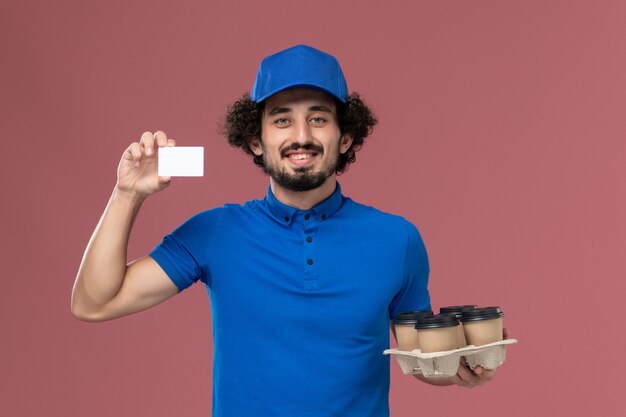 Vista frontal del mensajero masculino con gorra uniforme azul con tazas de café de entrega y tarjeta de plástico en sus manos en la pared rosa