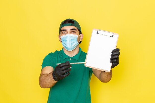 Vista frontal de mensajero masculino joven en camisa verde gorra verde anotando notas que muestran el bloc de notas sobre el fondo amarillo entregando el color del servicio