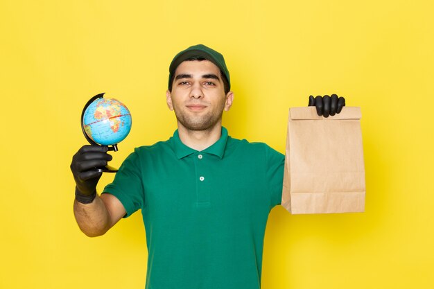 Vista frontal mensajero masculino joven en camisa verde gorra verde con paquete de entrega y pequeño globo en amarillo