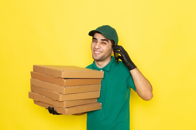 Vista frontal mensajero masculino joven en camisa verde gorra verde sosteniendo cajas de entrega y hablando por teléfono en amarillo