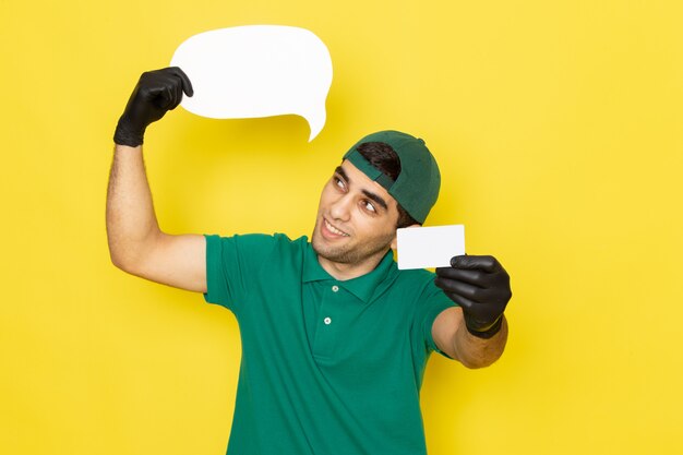 Vista frontal de mensajero masculino joven en camisa verde gorra verde con tarjeta blanca y cartel con sonrisa en amarillo