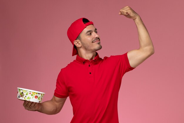 Vista frontal mensajero masculino joven en capa uniforme roja flexionando y sosteniendo el tazón de entrega sobre fondo rosa claro.