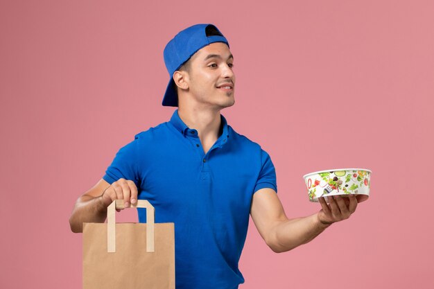 Vista frontal de mensajero masculino joven en uniforme azul y capa con paquete de entrega y tazón en sus manos en la pared rosa