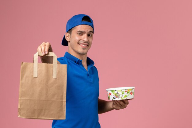 Vista frontal de mensajero masculino joven en uniforme azul y capa con paquete de entrega y tazón en sus manos en la pared rosa