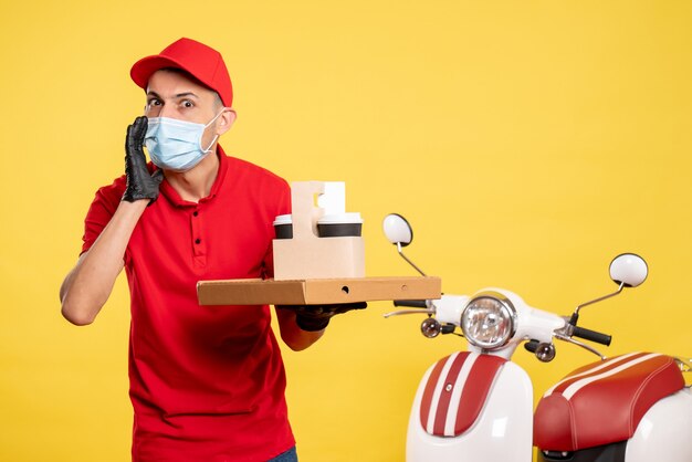 Vista frontal mensajero masculino en máscara con entrega de café y caja en el servicio de trabajo de color amarillo covid- virus uniforme de trabajo pandemia