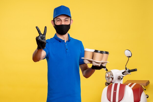 Vista frontal mensajero masculino en máscara negra sosteniendo café en amarillo trabajo de entrega uniforme de servicio de trabajo pandemia covid