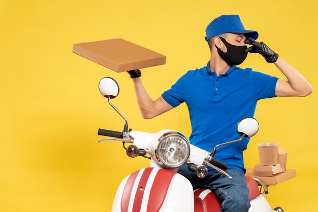 Vista frontal mensajero masculino en uniforme azul con caja de comida en amarillo virus pandemia covid- servicio de trabajo trabajo bicicleta