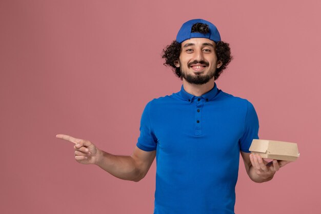 Vista frontal mensajero masculino en uniforme azul y capa sosteniendo un pequeño paquete de comida de entrega en la pared rosa uniforme trabajo de empresa de servicio de entrega masculino