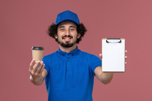 Vista frontal del mensajero masculino en uniforme azul y gorra con taza de café de entrega y bloc de notas en sus manos en la pared rosa