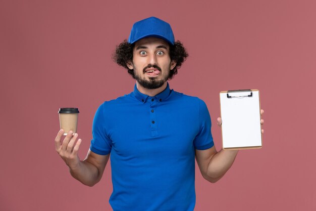 Vista frontal del mensajero masculino en uniforme azul y gorra con taza de café de entrega y bloc de notas en sus manos en la pared rosa