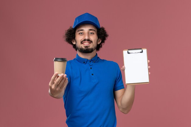 Vista frontal del mensajero masculino en uniforme azul y gorra con taza de café de entrega y bloc de notas en sus manos en la pared rosa