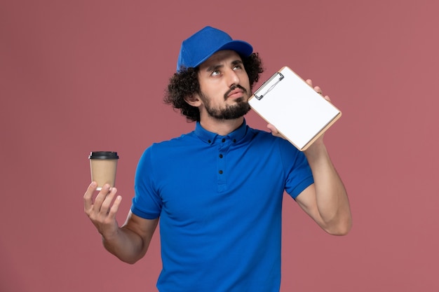 Vista frontal del mensajero masculino en uniforme azul y gorra con taza de café de entrega y bloc de notas en sus manos pensando en la pared rosa