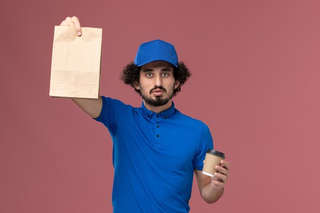 Vista frontal del mensajero masculino en uniforme azul y gorra con taza de café de entrega y paquete de comida en sus manos en la pared rosa