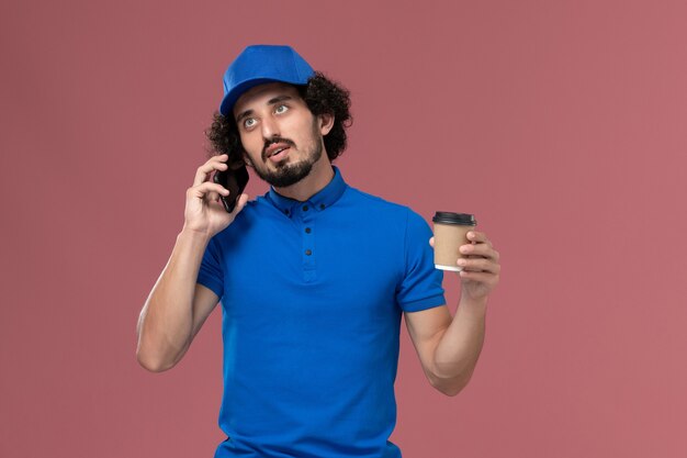 Vista frontal del mensajero masculino en uniforme azul y gorra con taza de café de entrega en sus manos hablando en la pared rosa