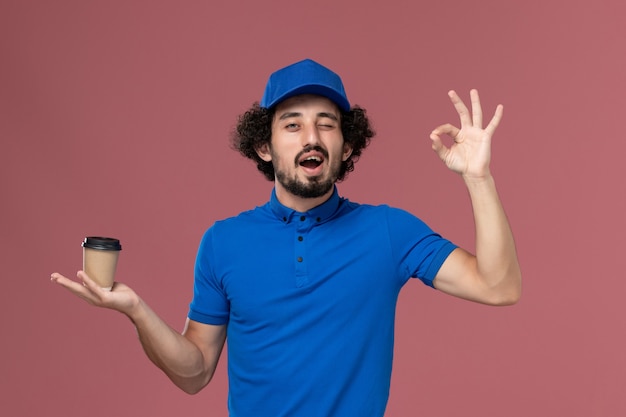 Vista frontal del mensajero masculino en uniforme azul y gorra con taza de café de entrega en sus manos en la pared rosa