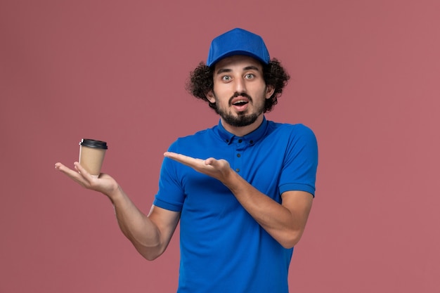 Vista frontal del mensajero masculino en uniforme azul y gorra con taza de café de entrega en sus manos en la pared rosa