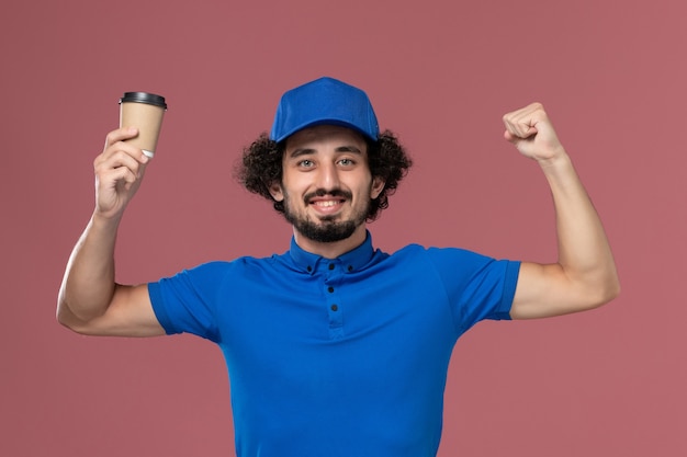 Vista frontal del mensajero masculino en uniforme azul y gorra con taza de café de entrega en sus manos en la pared rosa