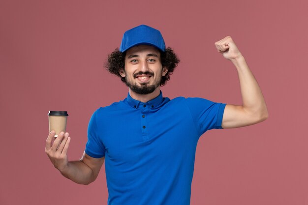 Vista frontal del mensajero masculino en uniforme azul y gorra con taza de café de entrega en sus manos regocijándose en la pared rosa