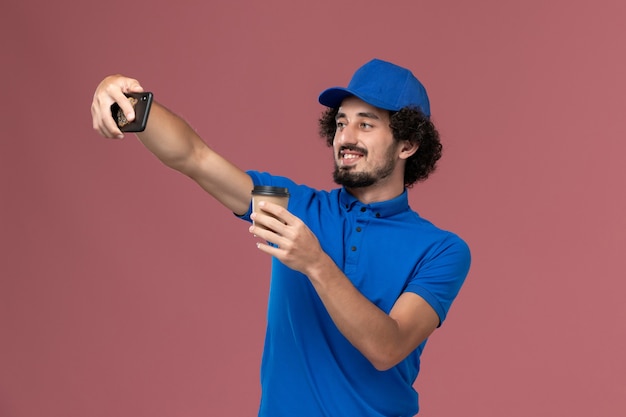 Vista frontal del mensajero masculino en uniforme azul y gorra con taza de café de entrega en sus manos y tomando una fotografía en la pared rosa